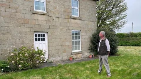 Martin Beal is looking at his house which is a Georgian stone built farmhouse with a white door and windows. He is a man, about 60 years of age, wearing a waist coat, white shirt and light coloured trousers.