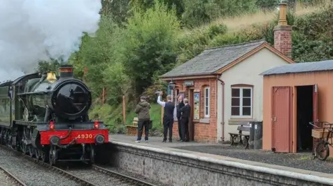A group of men on a platform waving towards a heritage train which is dark green with a red front. The men are standing in front of a small brick building. A pink shed is also on the platform with a bike with a basket on it. 