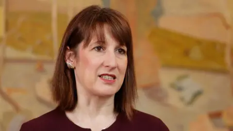 Rachel Reeves wearing a burgundy jumper. She is standing in front of an orange, yellow and gold background. 