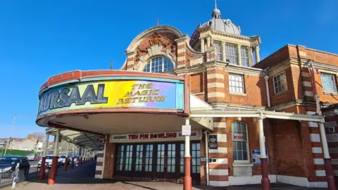 The Kursaal building in Southend-on-Sea. It has a brick front with a large tower and domed roof. There is a colourful curved sign at the front which says "Kursaal - the magic returns"