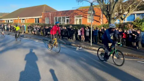 The three headteachers cycling along the road past a crowd of children on the pavement, who are smiling, waving and clapping as they pass them.
