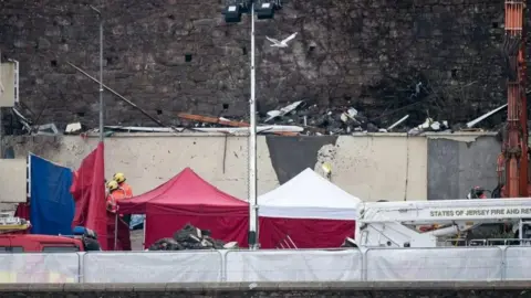 Firefighters are on the scene of the explosion, with rubble seen behind them. A States of Jersey Fire and Rescue Service vehicle can be seen.