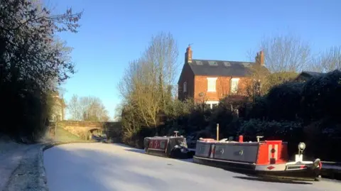 John Sadler The canal at Gonsall is completely frozen over in the winter and white with ice. Two boats are moored by the side. The sky is clear and light blue. There is a house in the background.