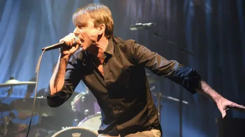 Brett Anderson of Suede performs on stage. He is wearing a black open-necked shirt and light-coloured trousers. He is holding a microphone in his right hand.