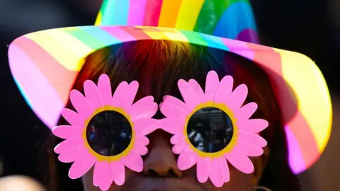 A woman wears a rainbow-coloured hat and pink-petal sunglaesses.