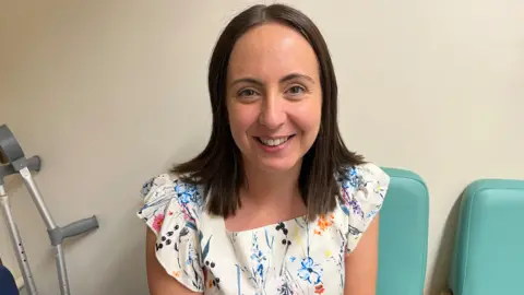 Ollie Conopo/BBC Kelly Roberts, who has brown hair and is wearing a white sleeveless top with flowers on, sitting on a chair. There is a light-coloured wall behind her.