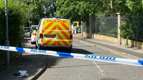 Police vehicles parked on a road behind a police cordon. The road is cordoned off with blue and white police tape. People in white suits can be seen in the distance.