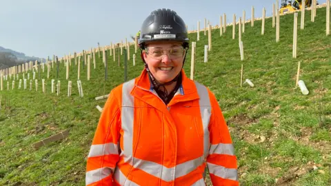 A woman wearing an orange hi vis jacket, construction goggles and a black construction helmet smiles as she stands on a sloping field with small saplings planted across it. The sky above is blue.