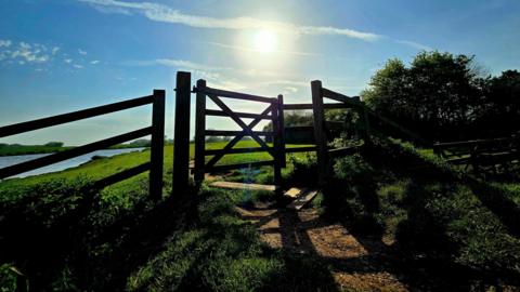 Blue sky with bright sun shining over a field with a gate in the foreground. 