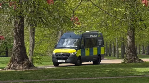 A yellow and blue police van is parked on a path among trees in Kensington Gardens