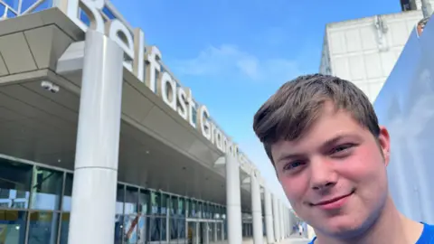 Luke is standing outside the new station. He was brown hair swept to the right side. He's wearing a blue top and smiling at the camera. The station has white pillars holding it up and white lettering saying Grand Central Station. The sky is blue and there aren't many clouds. 