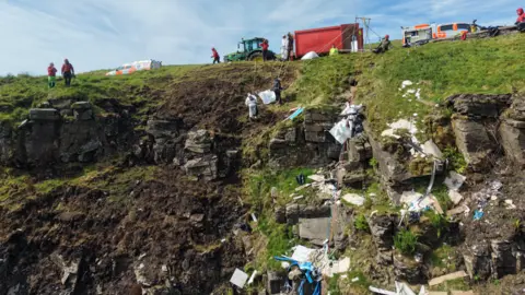 Central Beacons Mountain Rescue Team Close up drone image showing people cleaning up piles of rubbish on the side of a mountain. The volunteers are attached to ropes, with crews supporting them on the top of the hill.