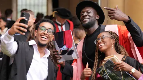 Stormzy is stood in the middle of two students. The man on the left is holding a phone taking a selfie. They are all looking towards the camera smiling. 