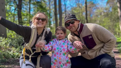 A man and woman smile at the camera while holding the hands of their daughter, a toddler who is stood between them. The couple kneel beside her in a forrest.