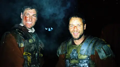 Andy Bowden standing next to Russell Crowe while filming in Bourne Woods in 1999. Both are smiling and dressed in Roman armour costumes, with fake blood and bruising on their faces.