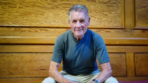 Sir Michael Palin in a t-shirt and trousers sat in a wood-panelled room. He is leaning on his knees and smiling for the camera.
