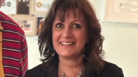 Surinder Hothi Surinder Hothi is seen from her shoulders up. She has shoulder-length brown hair and is smiling. There are framed pictures on a wall behind her.