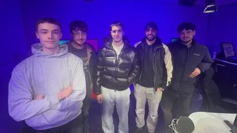 A photo of five young men stood in the Newsbeat studio. From left to right there is Reece who is wearing a grey hoodie with his arms crossed, then Anakin who is wearing a dark grey t-shirt and has glasses, then Kenan who is wearing grey joggers and a black puffer jacket, then Zeesham who is wearing jeans and a black jacket, and then Thaua who is wearing black jeans and a dark zipped up coat.