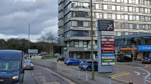 The outside of the Sunbury Cross Shopping Centre, in daylight, with traffic driving past.