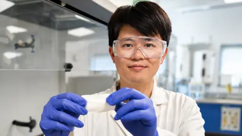 Dr Xiang Ding. He is pictured in a science lab wearing goggles, a white jacket and blue latex gloves. He is holding a plant-based wound dressing in front of the camera and is smiling.