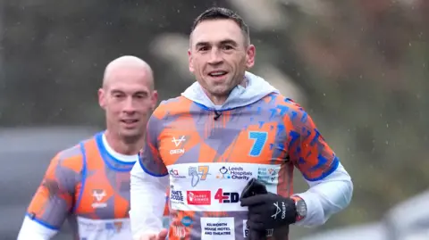 PA Media Kevin Sinfield running with a member of his support crew behind him. Sinfield is wearing an orange and blue top with the number 7 emlazoned on.