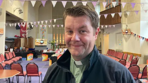 Wearing a green top with a dog collar and a black jacket, Rev. Patrick Webb stands in a church hung with flags.