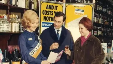 Barclaycard A photo from 1960s showing a female Barclaycard employee promoting the credit card in a shop lined with bottles of wine and spirits. There is a stand next to her saying "Barclaycard - Britain's first all-purpose credit card". She is showing a leaflet to a man and woman, with the woman holding a wicker basket.