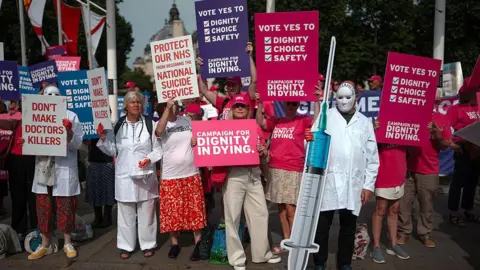 Supporters and opponents of assisted dying hold placards outside Parliament in June 2025. Supporters hold pink placards reading "Campaign for dignity in dying", while opponents, some wearing white medical coats and holding mock syringes, hold signs reading "Don't make doctors killers".