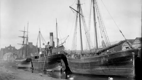 AHAG A black and white image of Annan harbour with three large ships in it