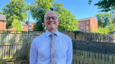 Jessica Bayley/BBC Image shows a bald man with round glasses smiling towards the camera. He is wearing a blue pinstriped shirt and a speckled tie, in the background are garden fences.