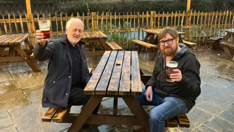 Two men sit at a picnic bench in a pub garden. They are both holding their pints of beer towards the camera. It is a wet day. The garden overlooks a river. One of the men has white hair and beard. The other has brown hair, a beard and glasses.