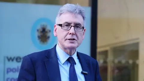 A man with grey hair, black rimmed glasses and wearing a navy blue suit is standing outside Lloyd House, the headquarters of West Midlands Police (WMP) in Birmingham.