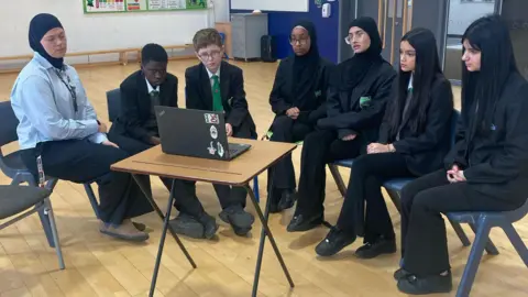 Six school pupils sit on chairs in a semi-circle with their teacher, watching something on a laptop 