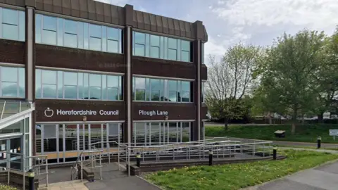 Google Herefordshire Council's offices in a modern building with large windows and a ramp with railings at the front. There is grass at the front of the building and an open space with trees to one side.