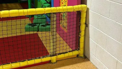 Ashley A children's softplay park is surrounded by yellow, foam covered posts and has colourful flooring. On the outside corner, a small brown rat can be seen lying on the floor between a post and the wall. 