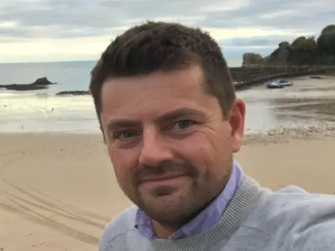 Mark Godfrey A man with brown hair wearing a lilac shirt and grey jumper smiles at the camera, behind him is a beach with a rock sea wall and yellow sand. 
