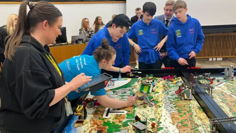 A bunch of people surrounding a table where a robot has had to take on various obstacles. The boys in blue tops are at one end while another person, a woman, is at the side, holding a tablet.
