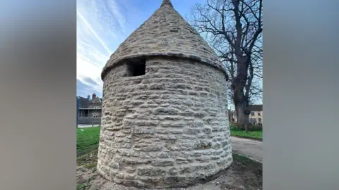 A stone-built round house with a domed roof. There is a small opening in a square window shape just beneath the roof. The structure has been cleaned and is positioned in a village green, with a large tree behind it and some residential housing. 