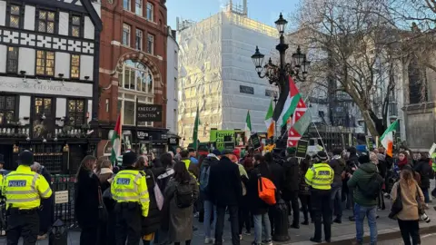 Supporters of Kneecap gather in a group outside court. A sign reads 'Kneecap Junction', there are police officers standing around the group. Members of the gathering are holding various signs and palestinian flags and tricolours. 
