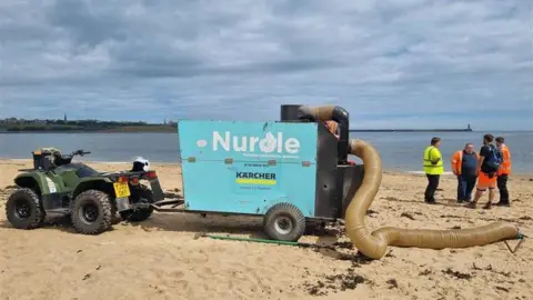 South Tyneside Council A blue trailer attached to a green quad bike on the sand. 'Nurdle' is written on the side of the trailer in white writing and the 'd' is in the shape of a shell. There is a long plastic tube coming out of the back of the trailer and is resting on the sand. There is seaweed on the sand and a group of four men in hi-vis standing to the right chatting. The sea is behind them and Tynemouth Pier can be seen in the background behind the men.