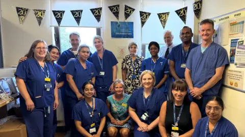 NHS Somerset A large group of medical staff - almost all of them in blue scrubs, line up for the camera. They are in two rows and above them is a line of bunting with the word "20" on it in white lettering on a dark background