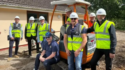 A group photo of members of Les Amis smiling, wearing hard hats and high visibility jackets. They are standing beside a piece of building equipment as they begin to break ground for their new site.
