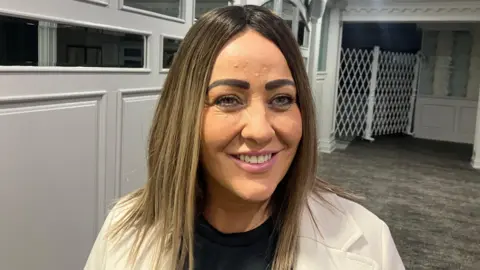 Angela smiling. She is wearing a black top and cream jacket. She has long light brown hair.