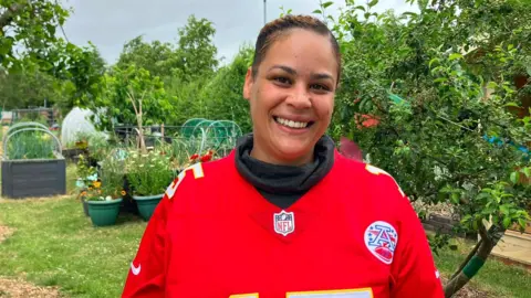 A woman in a red NFL jersey with grey polo neck underneath smiles in a garden, with pot plants, grass and raised beds behind her. The woman has dark brown hair tied back.