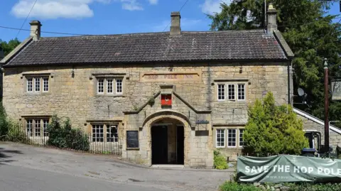 The Hop Pole pub in Limpley Stoke. It is an old two storey brick building with a pointed archway entrance and small, sunken sash windows. The roof is a dark slate and there are black metal railings separating the pub from the road, which is slanted on a hill. Outside the parking area to the right there is a green banner attached to a hedge that reads Save the Hope Pole