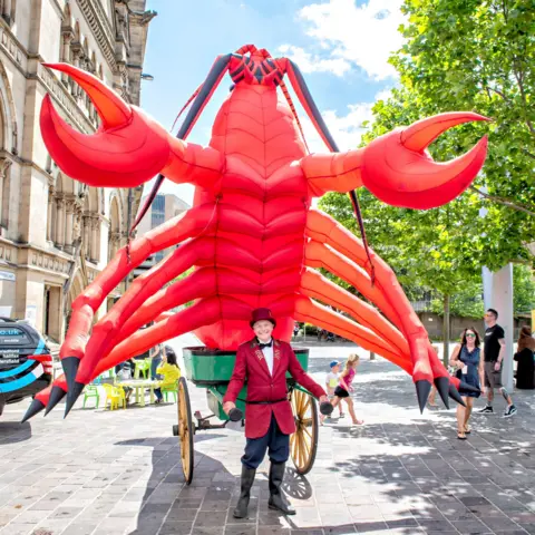 Tony Scales A man dressed in a red blazer, hat, blue trousers and black boots. He is stood in the street, pulling a green trailer behind him, which has a large, red, inflatable lobster in.