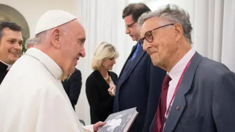 Pope Francis, dressed all in white, is holding the book and smiling as he talks to David Willey, who is also smiling.