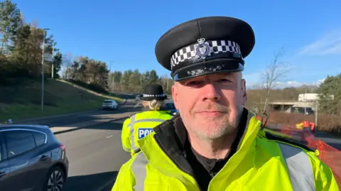 Dave looking into the camera and smiling slightly. He's wearing a yellow hi-vis jacket and a black peaked police officer's hat with the West Midlands Police badge on it. He's stood by the side of a road (Rampart Way in Telford) with cars visible to the left of the image. Over Dave's right shoulder there's another female police officer with her back to the camera - she's using a handheld camera to check cars' speed. By the side of the road there's grass and trees - and the sky is bright blue, with lots of sunshine.