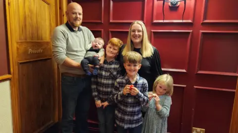 Family photo Nick and Amanda with their four children, three boys and a girl. The boys are wearing shirts and the girl wears a dress, and all are smiling in front of a red panelled wall.