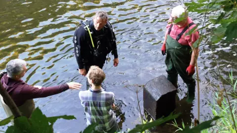 Supplied Gary Bankhead is standing in the water in a scuba diving equipment and suit. He has short grey hair. He is looking at a man with grey hair and red jumper, who is pointing to a large stone. A blonde man with a blue checkered shirt is standing nest to him, with his head turned in his direction. Another man in green overalls and red t-shirt and gloves is looking on. He has glasses and white hair.